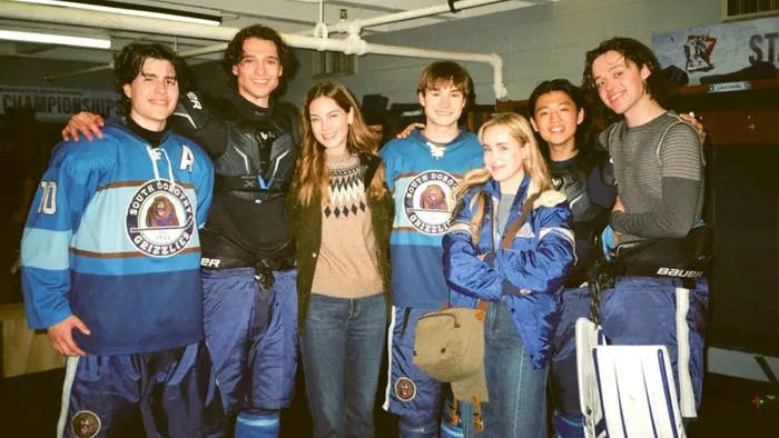 Group of young people in a locker room, with some wearing hockey gear. Others are dressed casually. Smiling and posing together for a photo