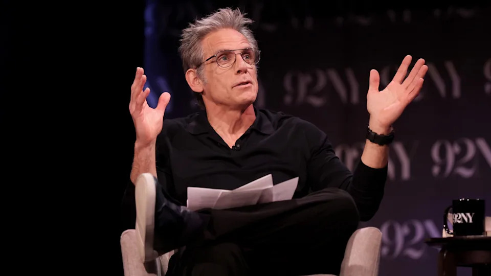 Ben Stiller speaking on stage, wearing glasses and a black shirt, gesturing with his hands.