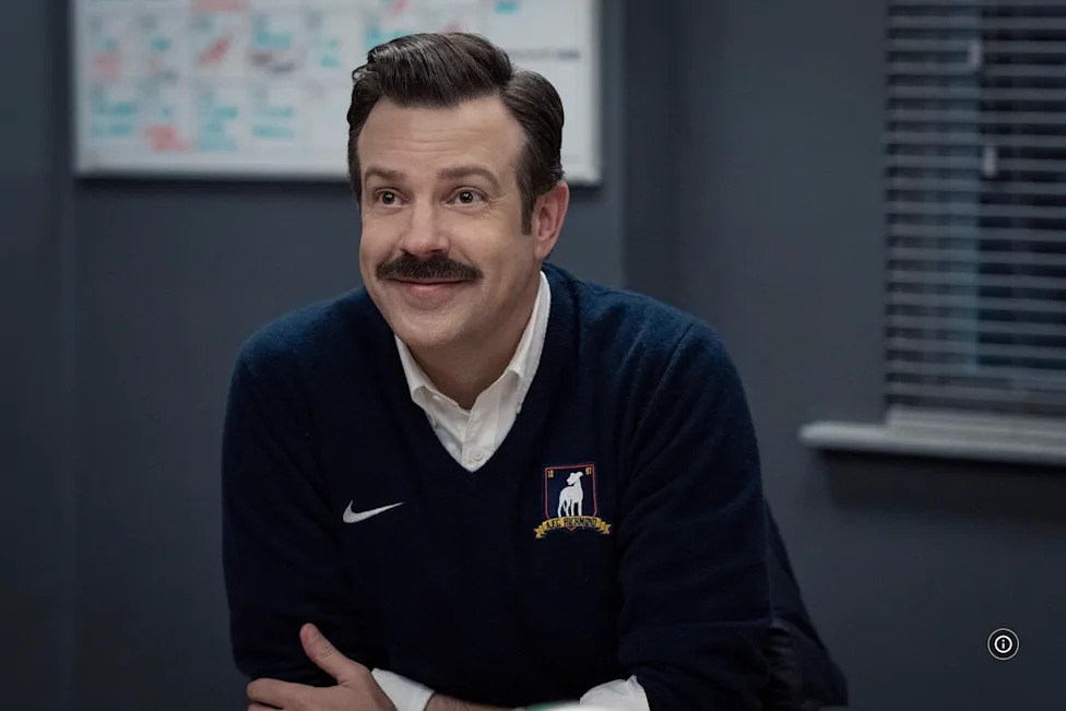 Man with a mustache smiling while sitting at a table, wearing a dark sweater with a logo.