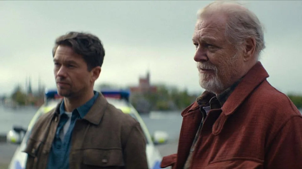 Jakob Oftebro stands next to an older man with a white beard, both looking ahead. A police car is behind them.