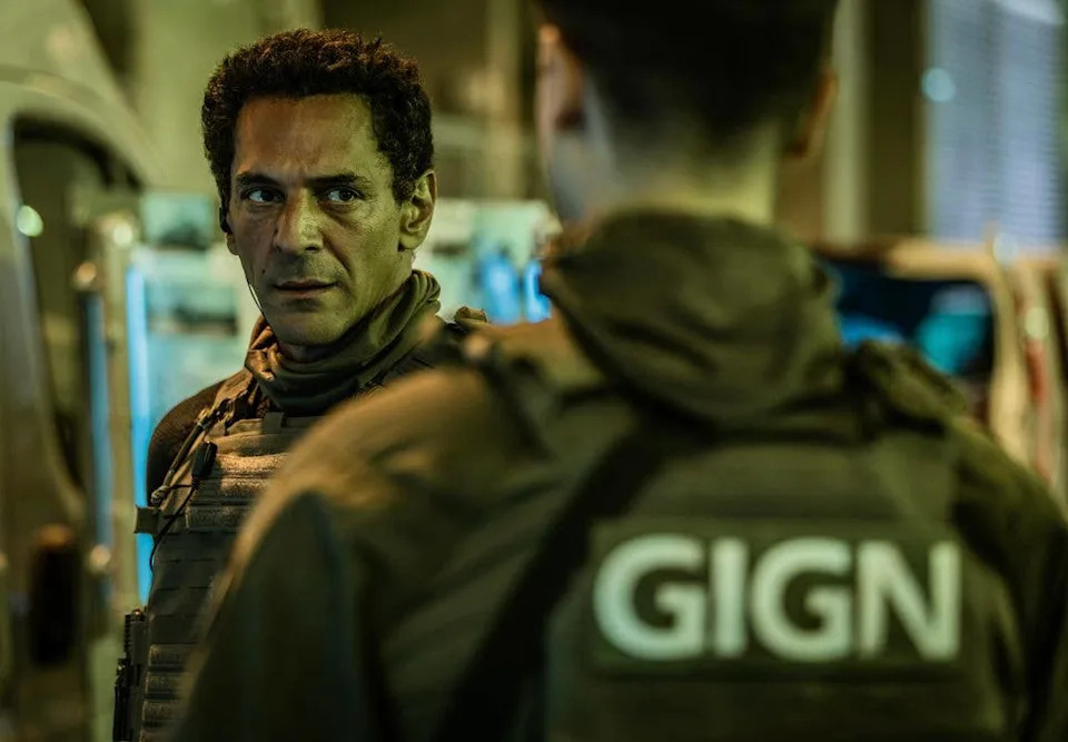 Tomer Sisley in tactical gear looks serious, facing a GIGN officer.