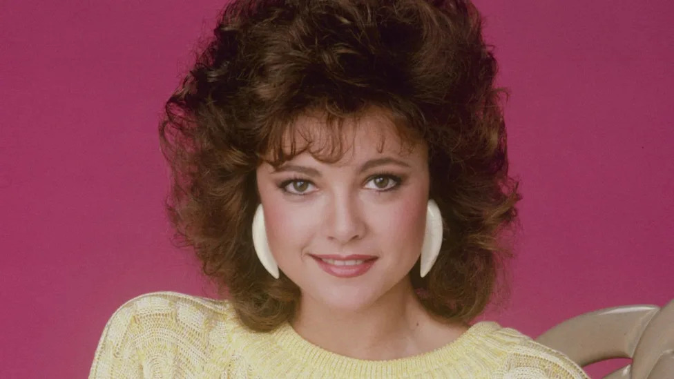 Emma Samms poses for a promotional portrait in front of a bright pink background, wearing a butter yellow jumper, dated 1985.