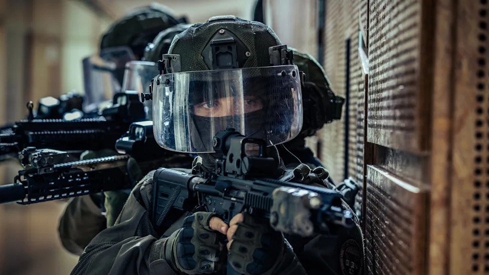Two tactical officers in full gear, one with a clear face shield, holding assault rifles.