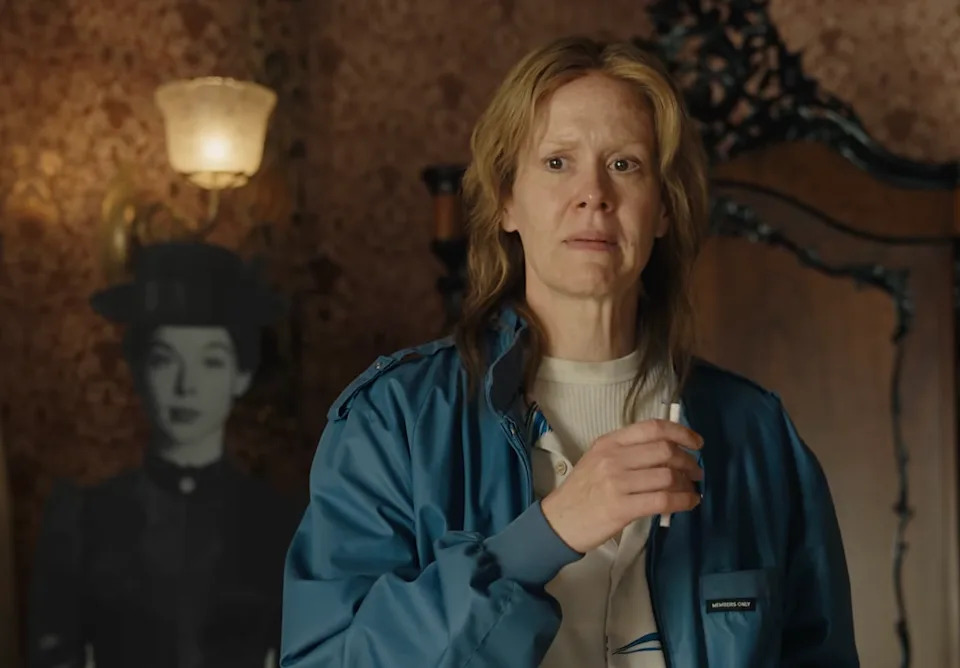 Sarah Paulson stands in a room, holding a cigarette, wearing a blue jacket.