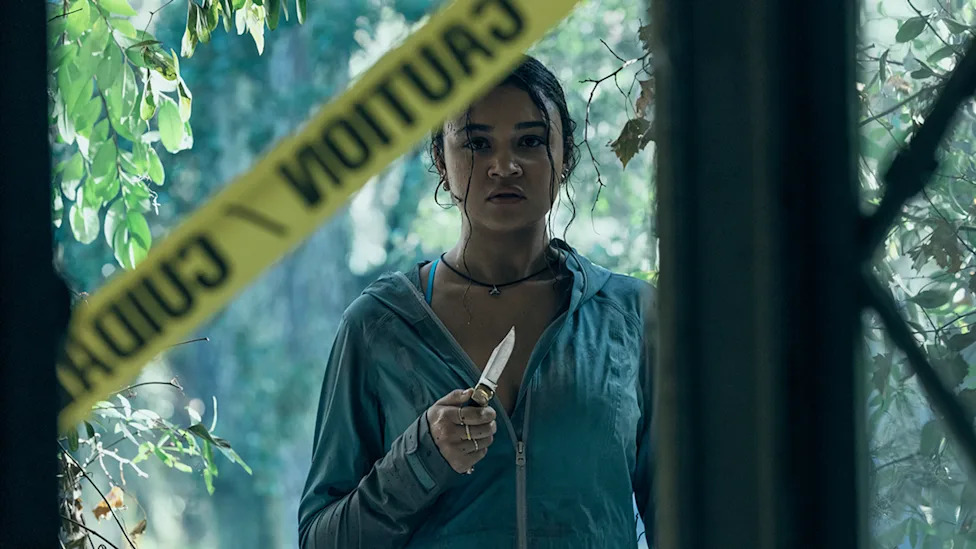 Madison Bailey stands in a doorway, holding a knife, with a 'Caution' tape behind her.