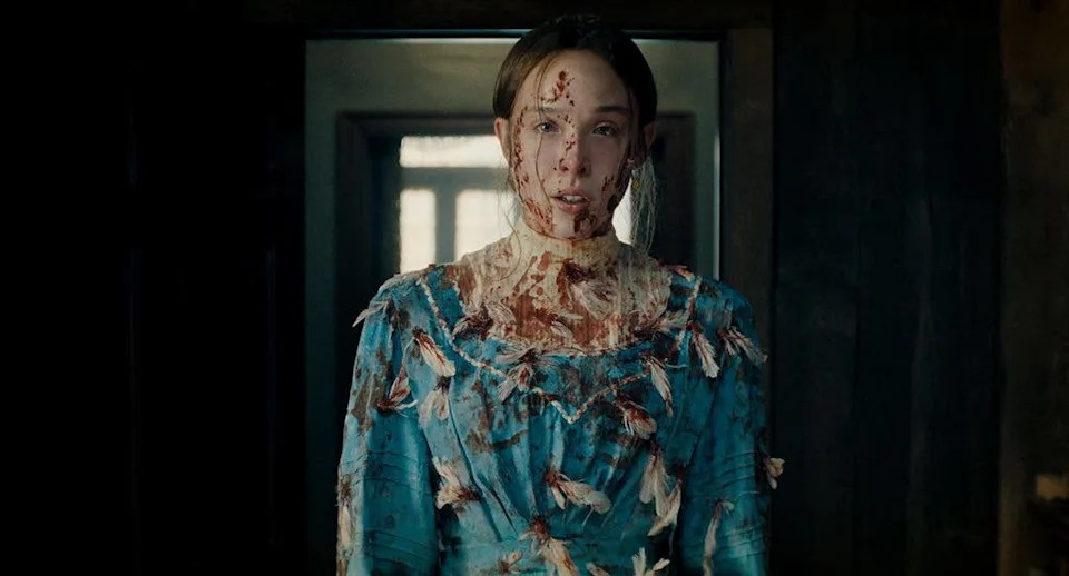 A woman in a blue dress with blood stains and feathers stands in a dark room.