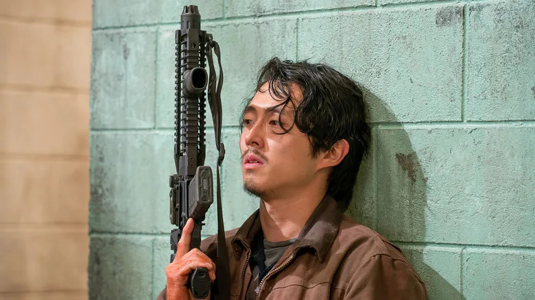 Glenn holding a rifle and leaning against a wall in "The Walking Dead"