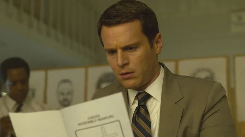 Jonathan Groff as Holden Ford standing in front of an investigation board looking at a manual in a scene from "Mindhunter."