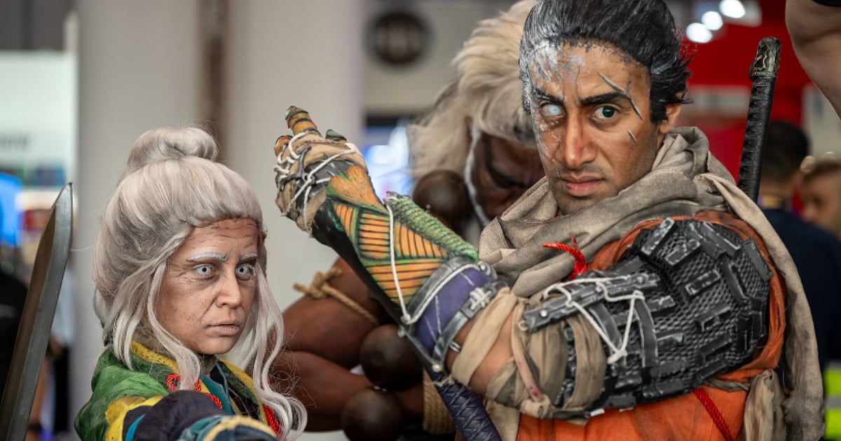 Two cosplayers in fantasy warrior costumes pose with weapons at a convention, with one wearing orange armor and the other dressed in green.