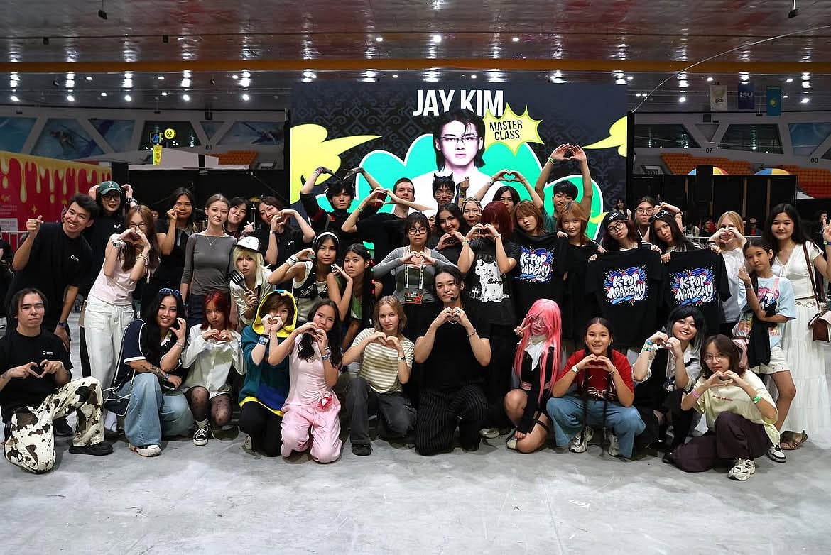 A large group photo of attendees posing with dance instructor Jay Kim following a K-Pop Academy master class. Participants of various ages are gathered in rows, many making hand-heart gestures toward the camera in front of a backdrop banner reading 'Jay Kim Master Class,' with several holding black K-Pop Academy t-shirts.