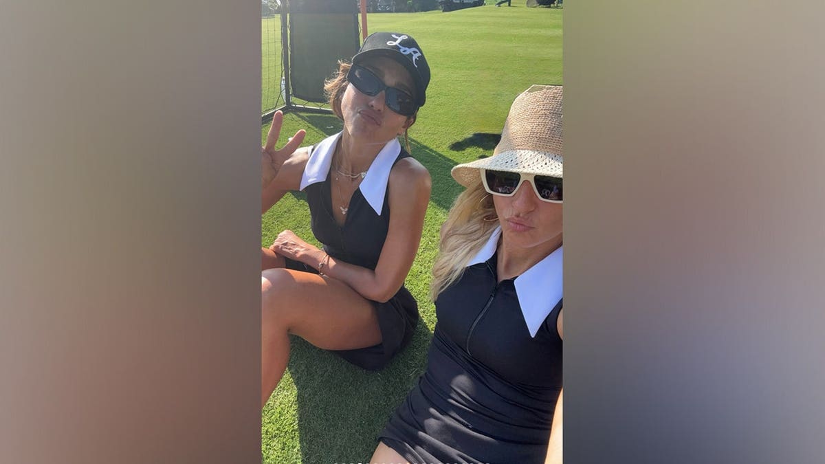 Jessica Alba and a friend pose together on a golf course wearing coordinating black sleeveless golf dresses with white collars.