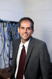 A man in a suit and tie stands in an office with a white brick wall and a desk with tangled cables in the background.