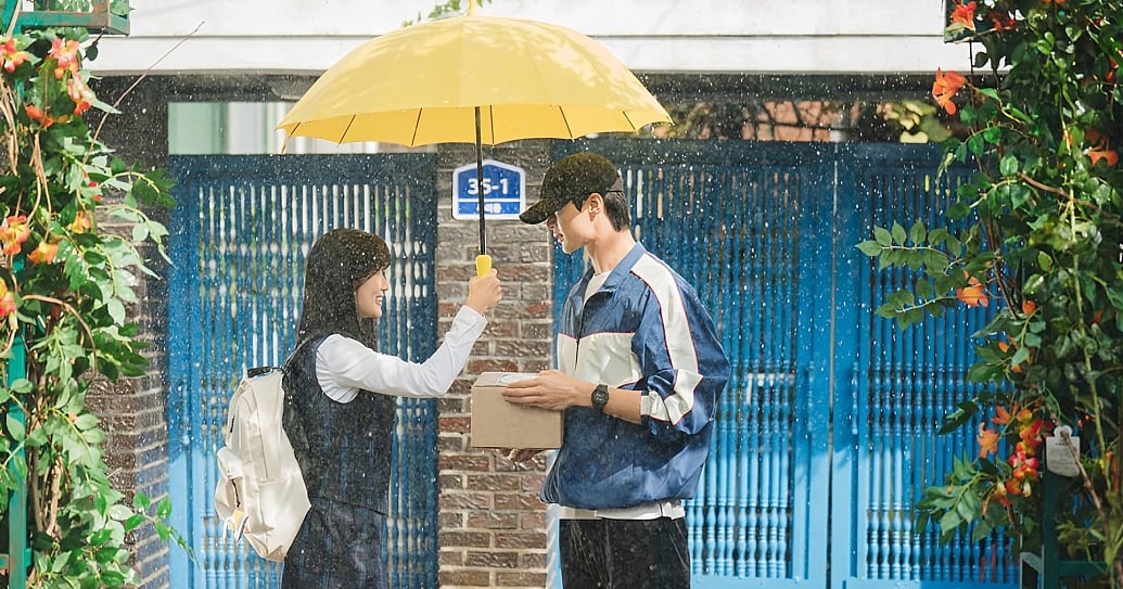 A scene from 'Lovely Runner' set in the pouring rain in front of a blue house gate framed by orange flowers. A high school girl wearing a uniform and white backpack holds a bright yellow umbrella over a young man wearing a black cap and blue windbreaker who is holding a cardboard parcel