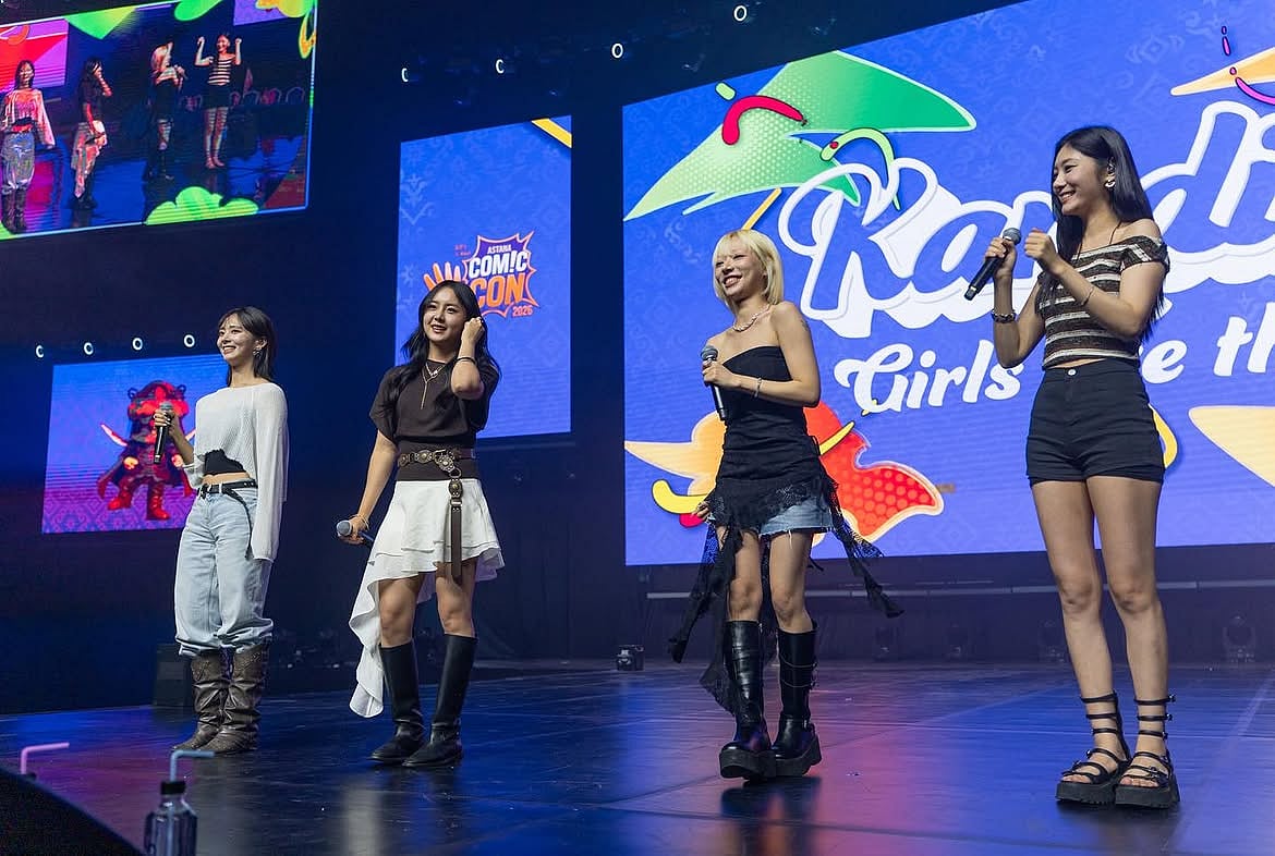 Four members of a girl group holding microphones on a stage during a performance at Astana Comic Con. They stand side-by-side wearing stylish casual outfits—including light jeans, asymmetric skirts, black platform boots, and striped tops—set against large background LED screens displaying colorful event graphics.
