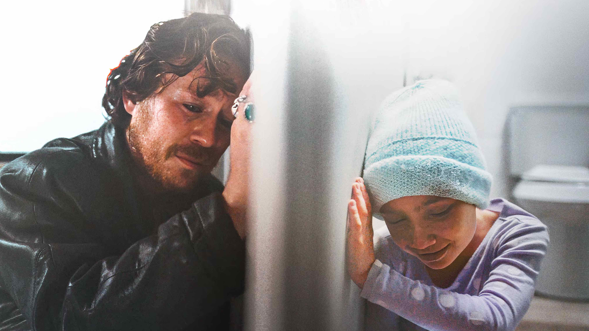 Robert Malcolm Cumming sits beside a young girl wearing a knit cap as they lean against a wall.