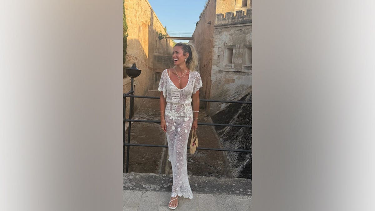 Kristin Cavallari wears a white crochet cover-up over a white swimsuit while posing beside a stone canal lined with historic buildings during her European vacation.