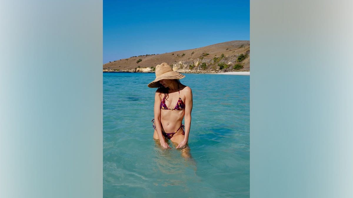 Michelle Monaghan looks down while standing in turquoise water in a colorful string bikini and wide-brimmed sun hat.