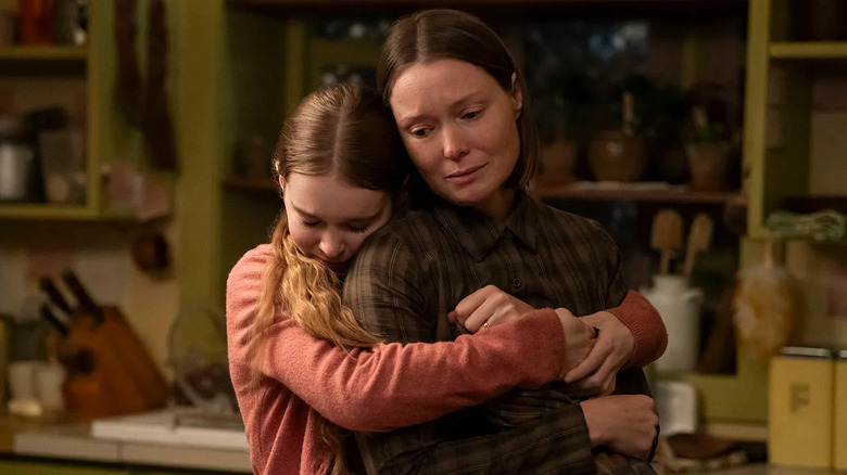 A young girl with brown hair is wearing a pink sweater and is hugging her mother in the kitchen. Her mother is wearing a plaid shirt and her brown hair is cut into a short brown bob.
