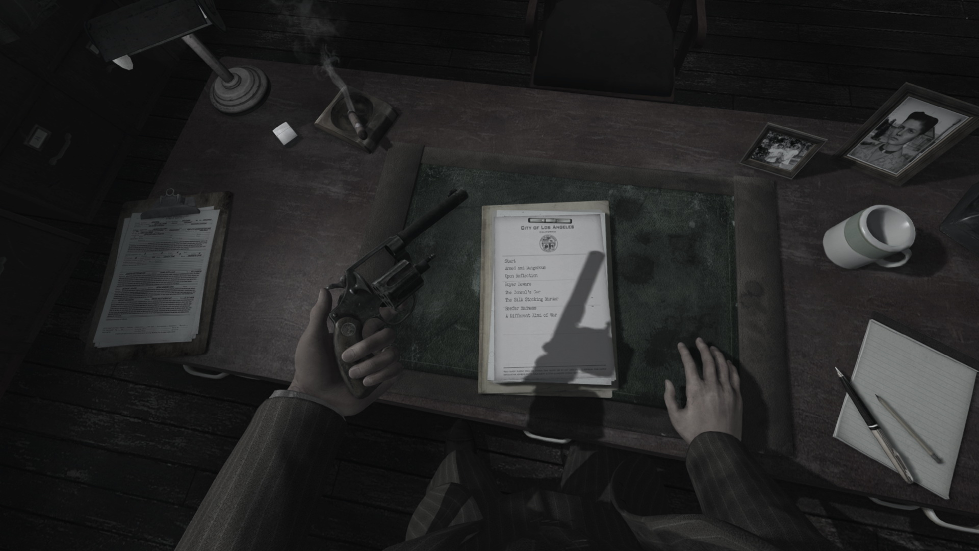 A first-person view from inside L.A. Noire: The VR Case Files, looking down at the player's own hands on a detective's desk, one holding a revolver, beside a City of Los Angeles case list, a smoking ashtray and a coffee mug