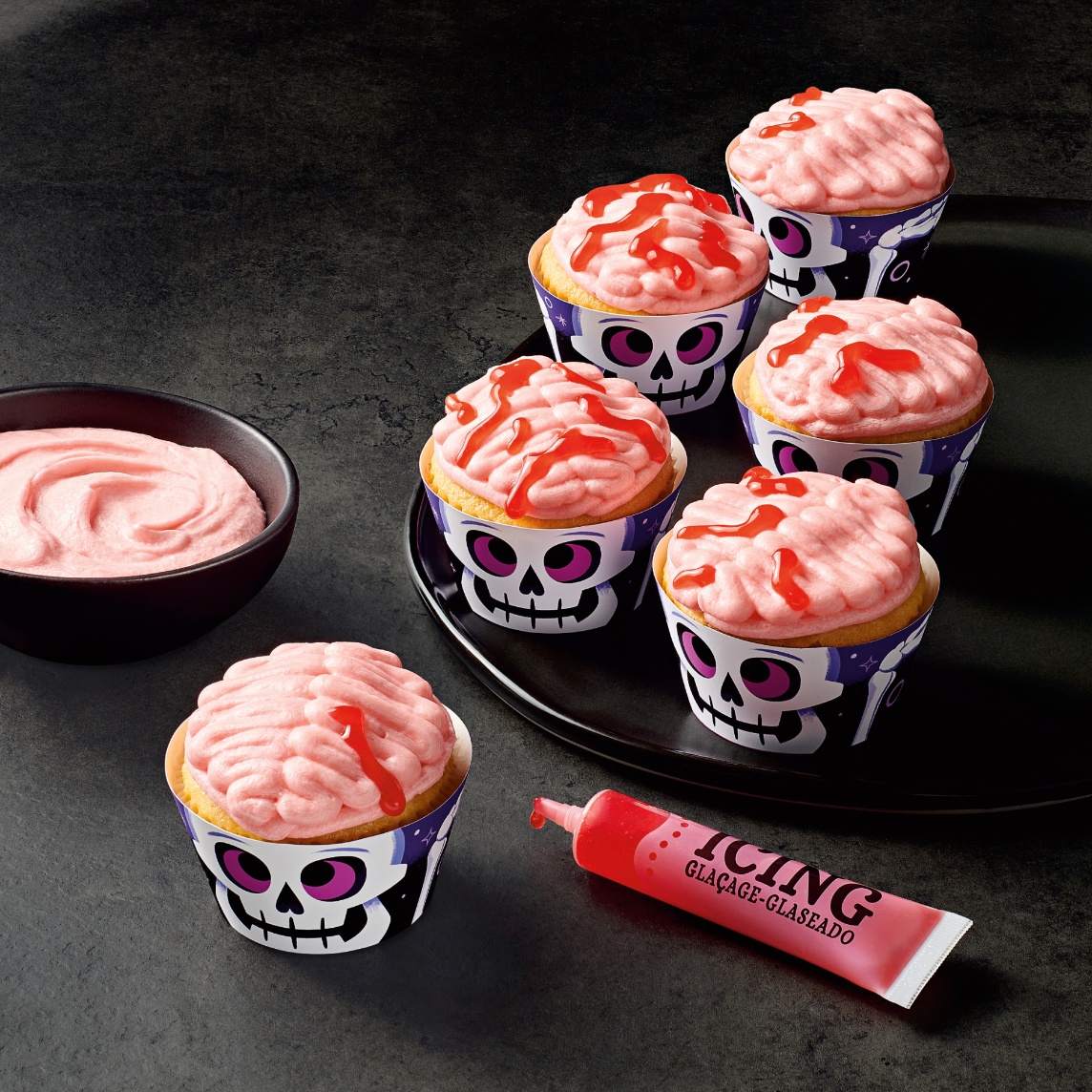 Brain-shaped cupcake decorating kit featuring frosted cupcakes with skull wrappers, pink icing and red decorating gel.