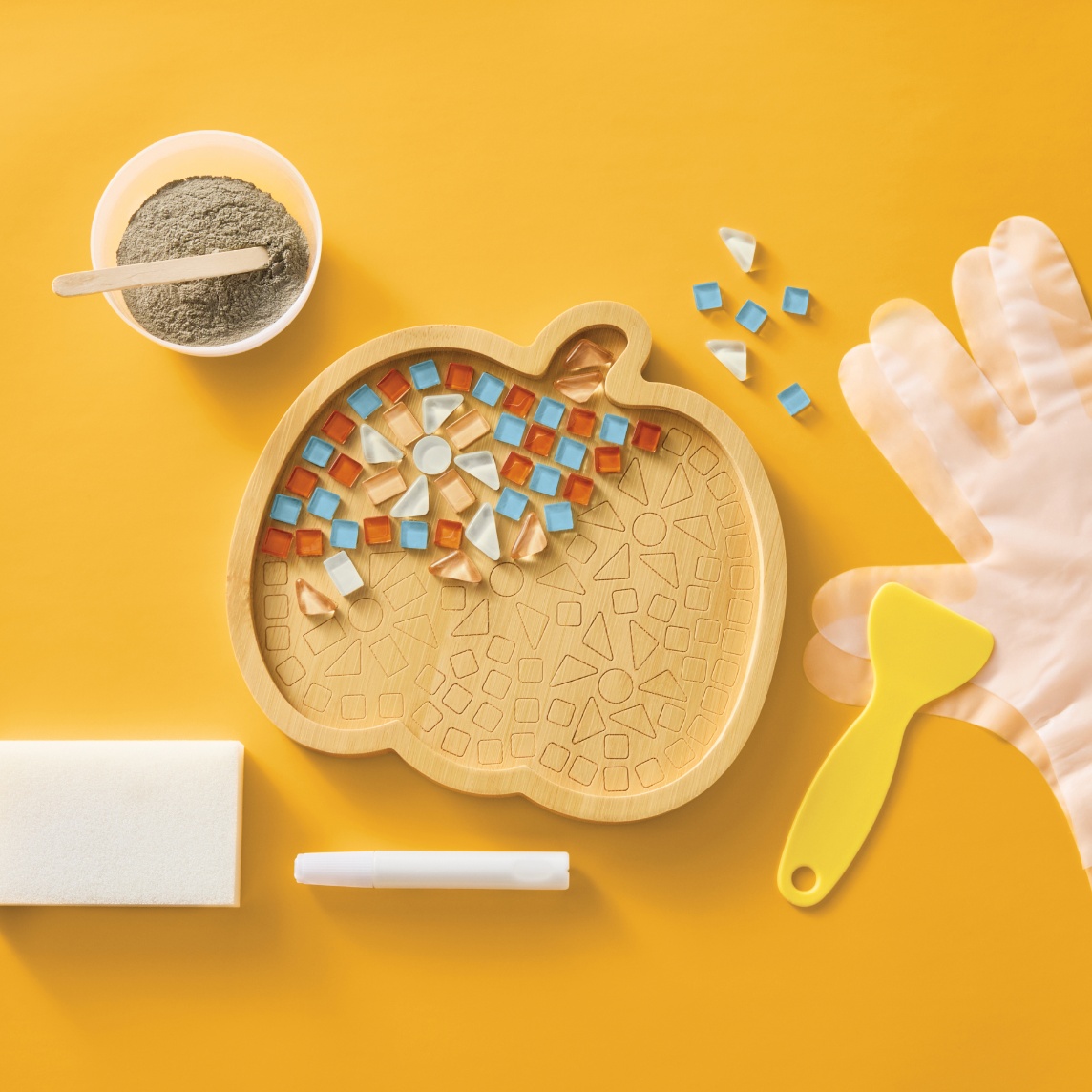 Pumpkin-shaped mosaic art kit with colorful tiles, adhesive, craft tools and gloves on a yellow background