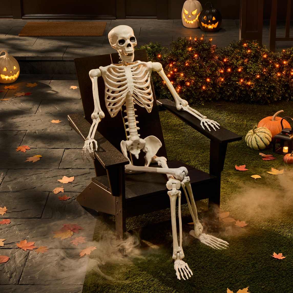 Poseable skeleton decoration relaxing in a black Adirondack chair surrounded by glowing Halloween decor, pumpkins, fall leaves and fog.