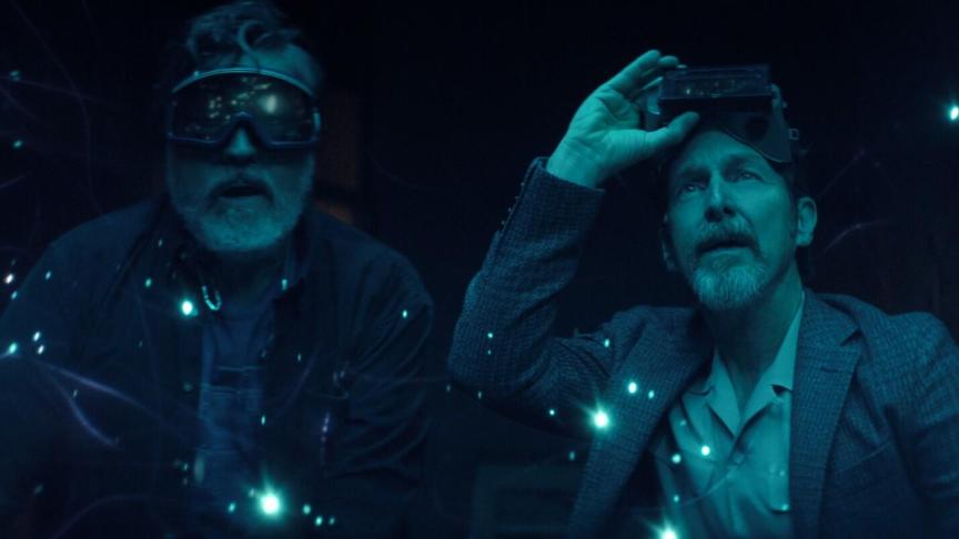 In a still from The Boroughs, a man in goggles on his face stands next to a man with goggles pulled off, looking in wonder and blue sparkling lights.