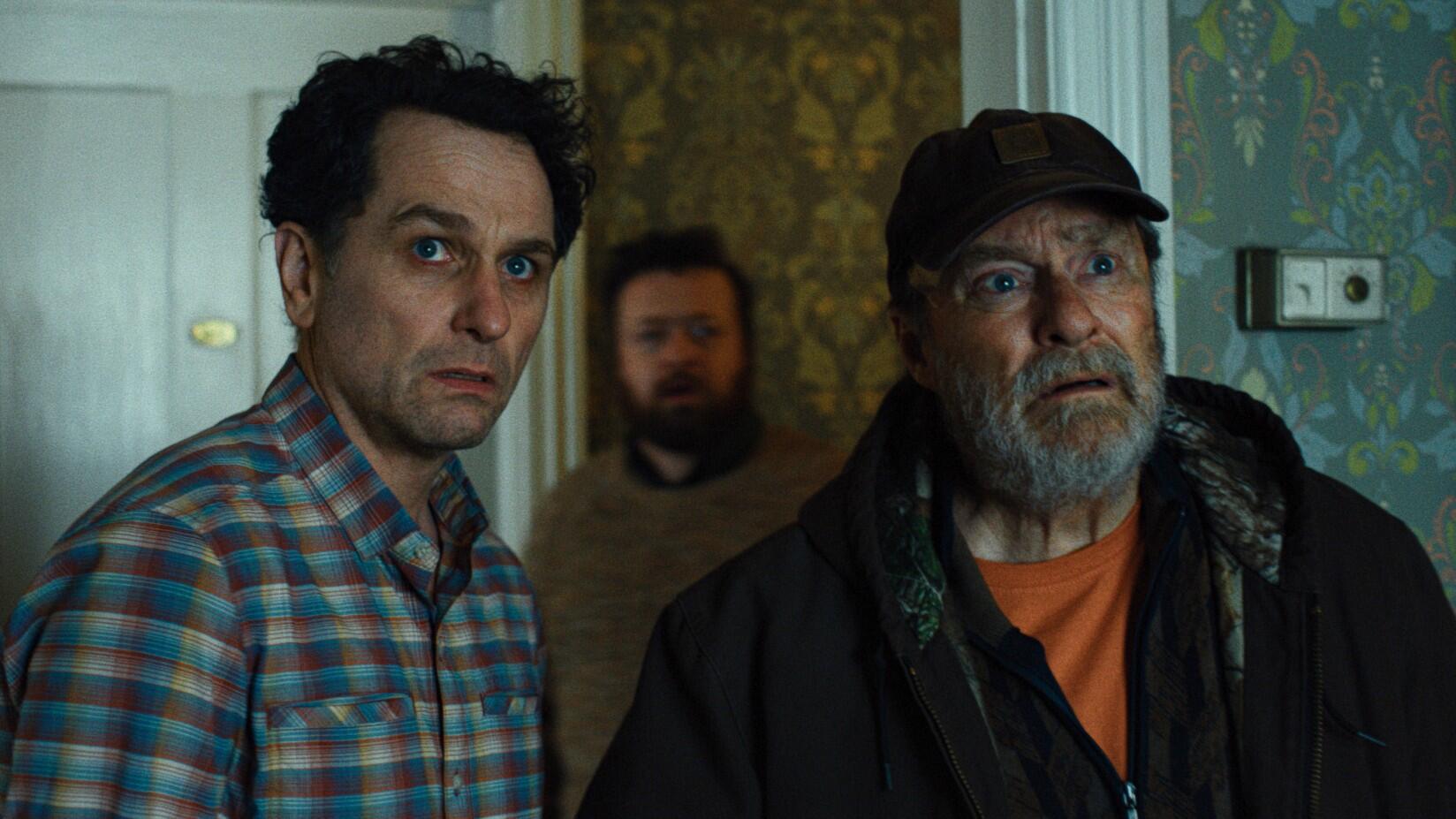Production still from Widow's Bay showing three white men looking scared in a wallpapered room.