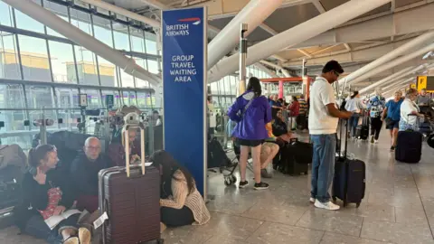 Jonathan Brady/PA Wire Passengers queue and sit on the floor at an airport. A large British Airways banner with the words Group Travel Waiting Area can be seen behind them.