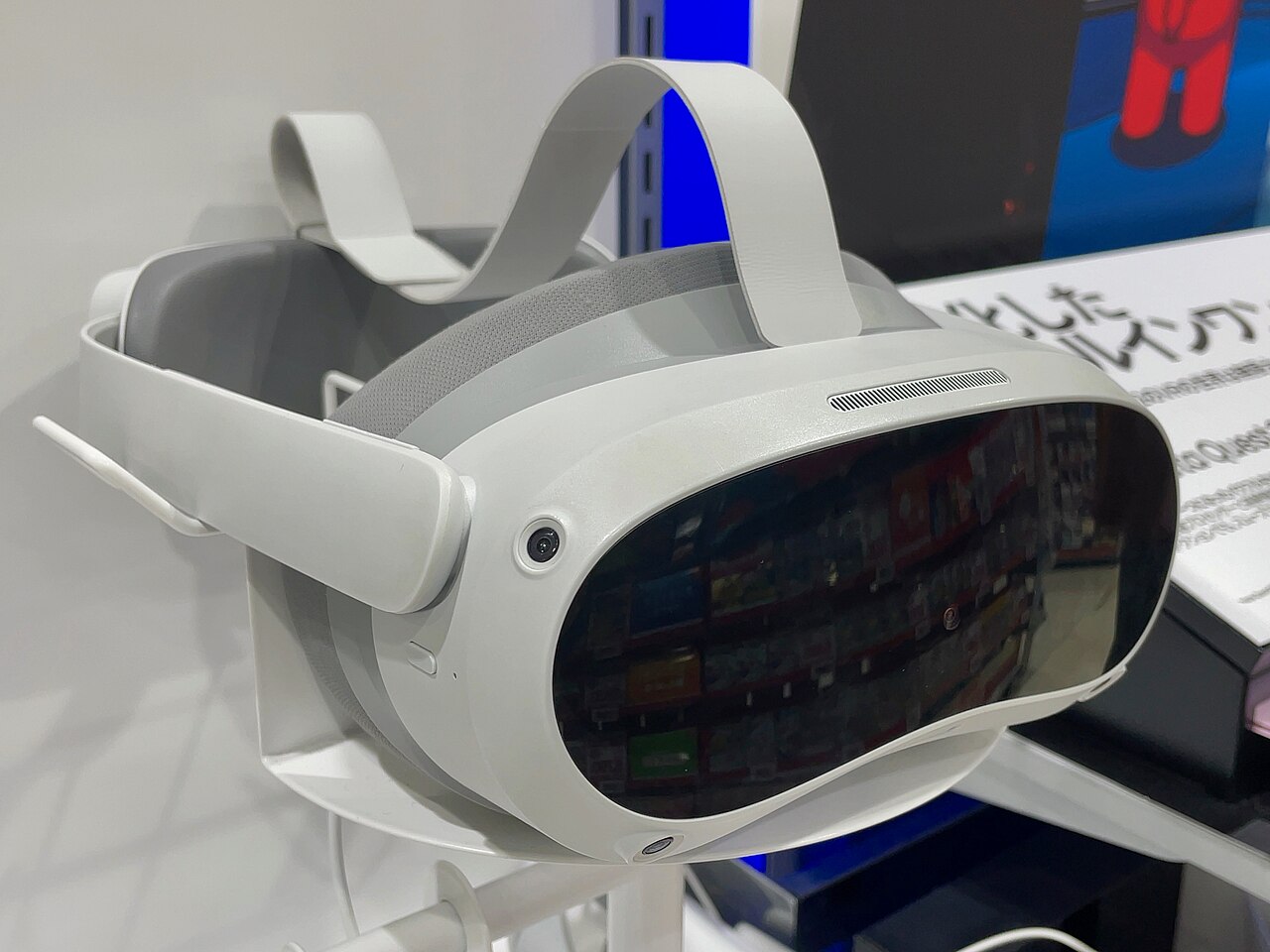 A white Pico 4 headset on a clear display stand in a retail store, front three-quarter view with the visor reflecting shop shelving