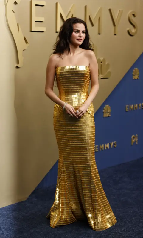 EPA Selena Gomez in a gold dress in front of a sign saying "Emmys"