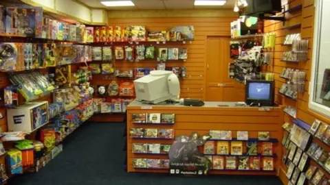 Dream Consoles an old picture of a videogame and gaming shop with a retro computer and tv, the walls are covered in gaming memorabvilluie ancd game cases.
