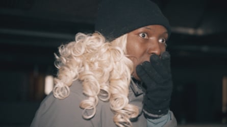 A head and shoulders side view of a Nigerian woman wearing a black woolly hat and white blond curls holding a black glove up to her mouth – with eyes wide open looking off to right of camera