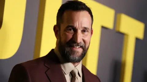 Getty Images Noah Wyle at The Pitt Los Angeles premiere, wearing a brown suit and beige shirt and tie