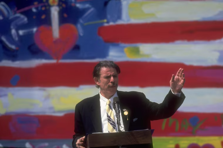 Peter Max gestures with one hand while standing at a lectern in front of a large scale oil painting.