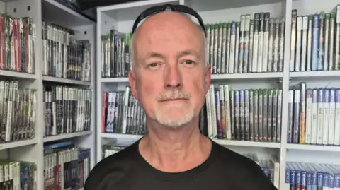 BBC An older man wearing a black t-shirt looking at the camera, he is in front of a shelf of game cases.