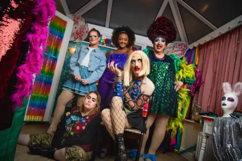 BBC/Hat Trick Productions A promotional shot for Smoggie Queens which shows three men in drag posing in a conservatory alongside two women.  In the foreground, one person sits on a chair wearing a blonde wig, fishnet tights, a patterned blue top and black lace-up boots, while another sits on the floor in camouflage shorts, black boots and a burgundy vest with badges. Behind them, three people stand wearing a denim outfit, a purple dress, and a green sequined dress with bright feathered sleeves. The room contains rainbow curtains, pink feather boas, costume accessories, and a white mannequin head wearing bunny ears on a small table to the right.