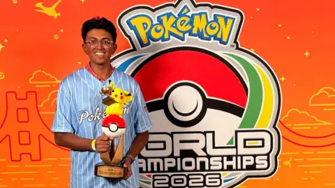 Supplied A teenage boy stands in front of a bright orange Pokémon World Championships 2026 backdrop, holding a trophy topped with Pikachu and wearing a light blue striped Pokémon shirt.