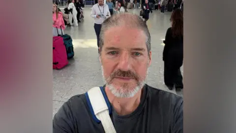 Charlie Adlard A man with light brown and grey hair an a beard stands in a busy airport. He is taking a selfie. He has a white and blue bag strap across one shoulder. Dozens of people can be seen behind him.