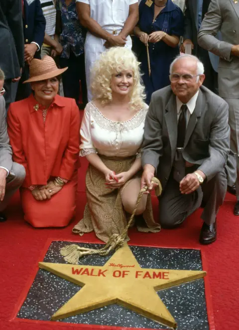 Getty Images Dolly Parton in 1984 kneeling and smiling on a red carpet in front of a large gold star saying "Hollywood Walk of Fame"