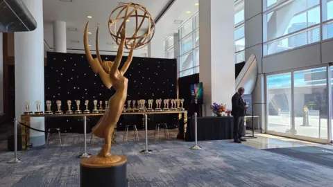 A large Emmy statue marks an entrance to a building. A table sits in the background with dozens of small gold trophies