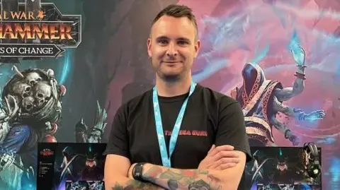 Supplied A man wearing a black tshirt with a blue lnayard standing in front of a video game poster smilintg at the camera