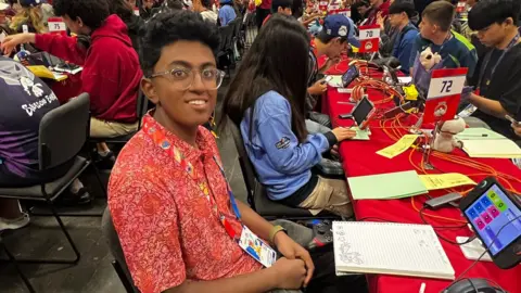 Supplied Rows of players sit at long red‑covered tables in a busy gaming hall, with numbered signs, screens and Pokémon‑themed banners, while the teenage boy at table 72 wears a red patterned shirt and lanyard, with a notebook, headphones and device in front of them.