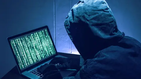 Getty Images A man wearing a black hoodie and black gloves has his back to the camera. His hood is up and you cannot see his face. His in a dark room and is working on a laptop which has lines of green numbers and letters on the screen.