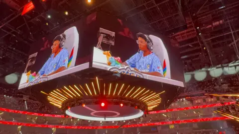 Supplied A large arena displays a gamer on a central hanging screen, wearing a blue striped Pokémon shirt and headset, with Charizard and Venusaur plush toys on the desk, surrounded by bright red and yellow lighting and a crowd of spectators.