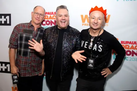 Santiago Felipe/Getty Images Three people standing in front of an event backdrop with visible logos and text including “RuPaul’s DragCon NYC 2018”, “WOW Presents Plus” and “#1”. The person in the centre wears a dark sequined jacket and raises both hands. The person on the left wears a plaid shirt and a black event pass on a lanyard. The person on the right wears a black sequined top with “VEGAS” printed on the front, a red paper crown, glasses hanging from a cord, and an “All Access” event pass.
