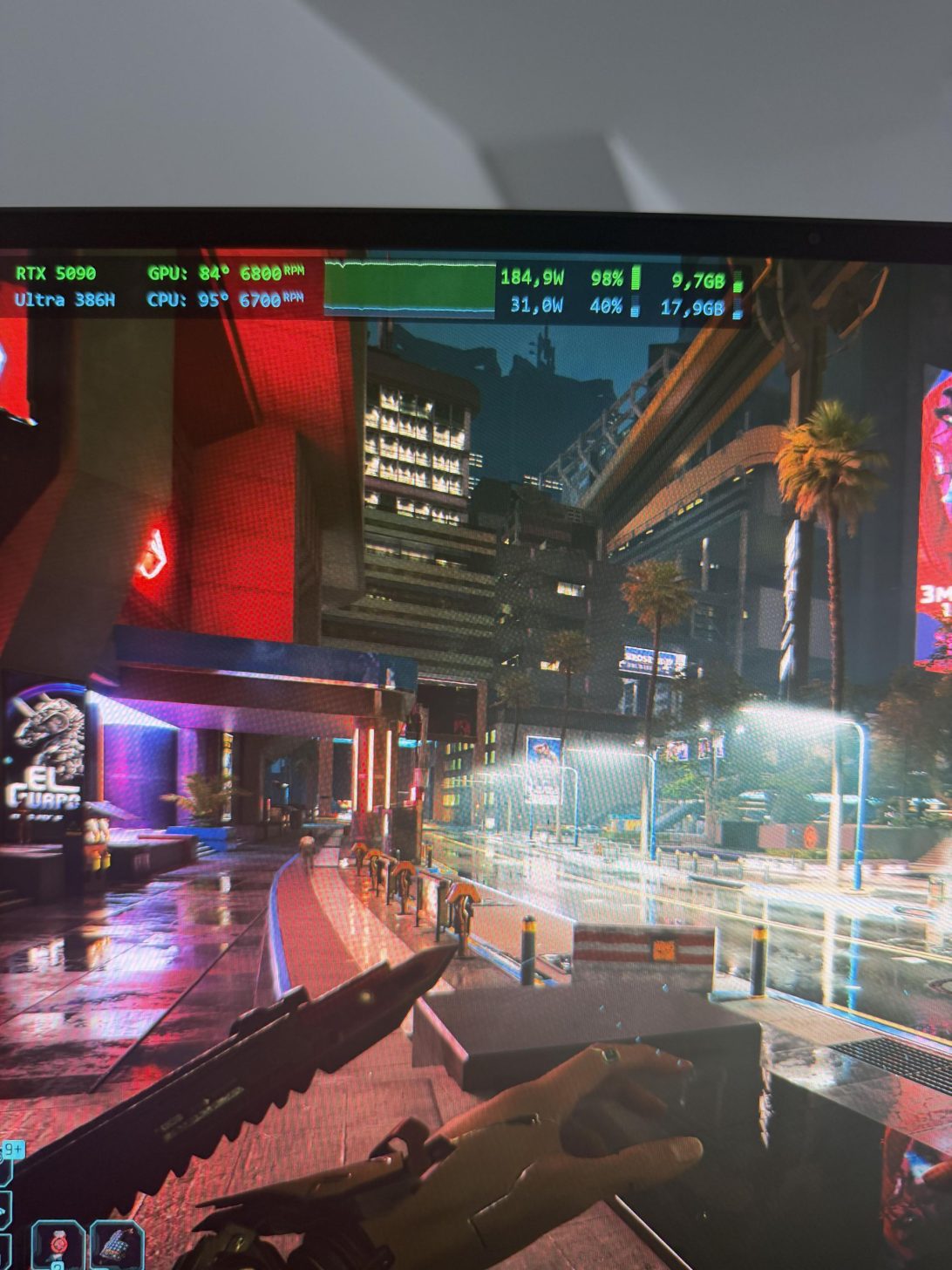 A gameplay scene from 'Cyberpunk 2077' shows a character holding a knife in a neon-lit city street, with on-screen performance metrics indicating 'RTX 5090,' GPU temperature '84°', and CPU temperature '95°'.
