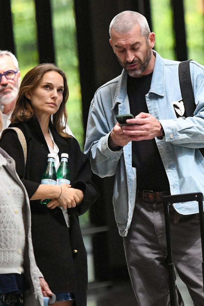 Natalie Portman and Tanguy Destable photographed outside. He is holding his phone in hand and looking down at the screen while she looks off to the right.