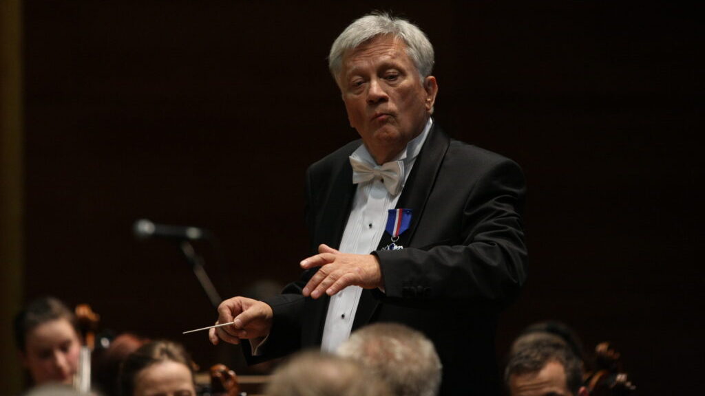 Cavatina Philharmonic Orchestra: An older conductor, Andrzej Kucybała, with gray hair and a white bow tie leads the orchestra, holding a baton and wearing a distinguished medal.