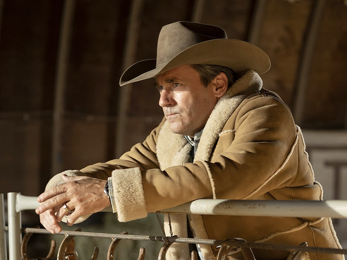 Jon Hamm wearing a cowboy hat and brown shearling coat, leaning on a railing in a dimly lit setting.